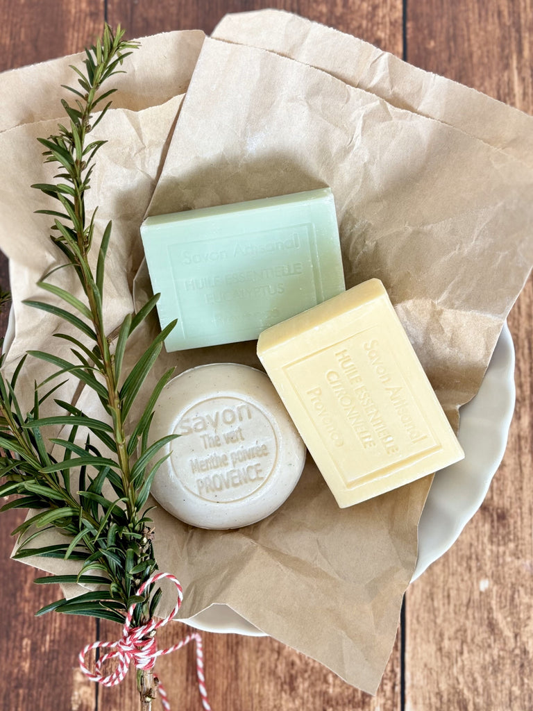 Three soap bars on crumpled paper in a white bowl with a green sprig tied with twine.