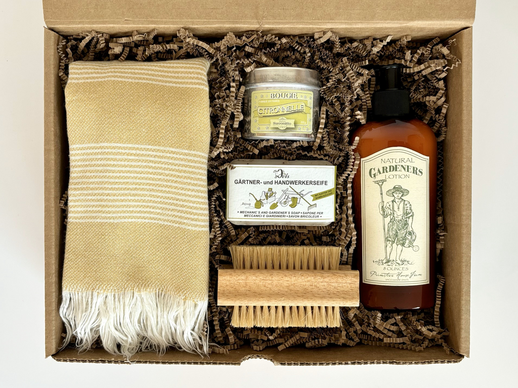 Top view of a gift box with personal care for gardeners, including a nail brush, a striped towel, a soap bar, a brown bottle of lotion, and a candle tin.