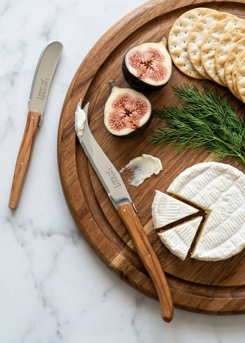 A wooden-handled butter spreader and a cheese knife and on a round wooden serving board with crackers, figs, brie cheese, and dill on a white marble surface. 