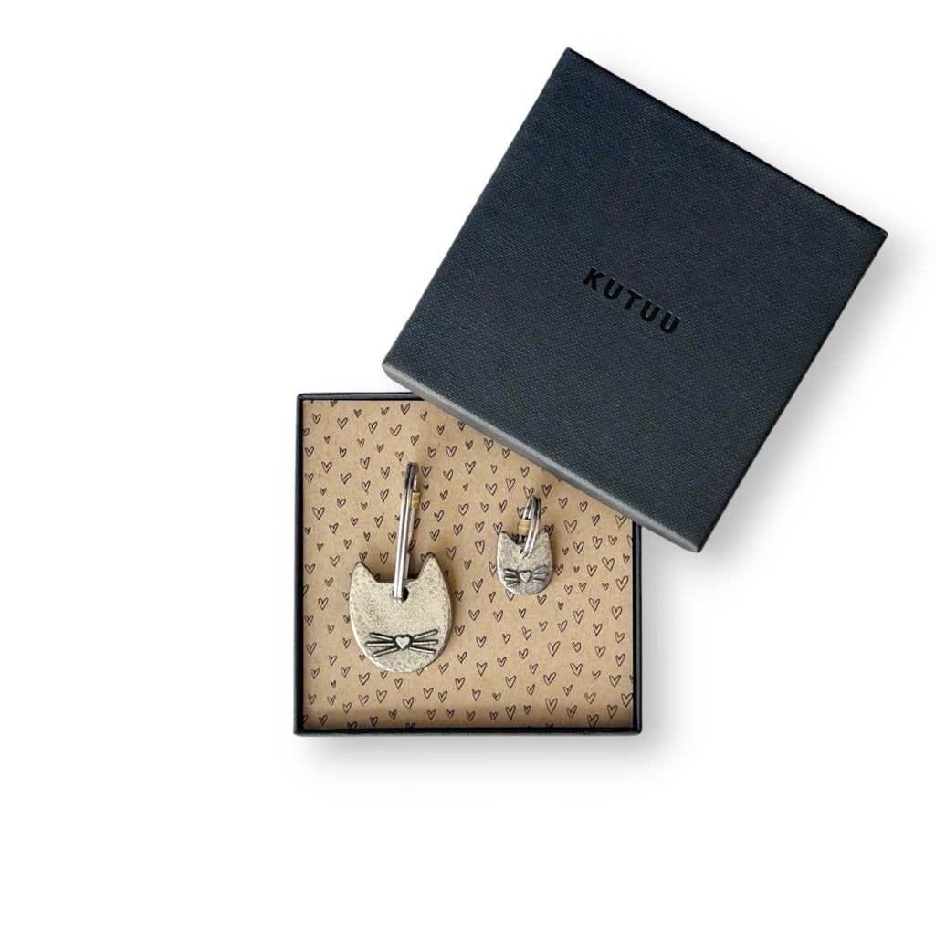 Gift box with two cat-shaped pewter keychain and charm on a heart-patterned lining, with "KUTUU" printed on the lid.
