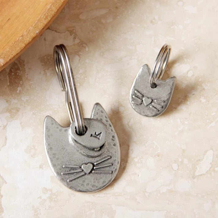 Two cat-shaped pewter keychain and charm with embossed heart noses and whiskers on a textured surface.