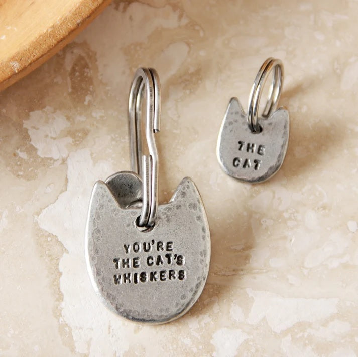 Two cat-shaped pewter keychain and charm with engraved text on a textured surface.