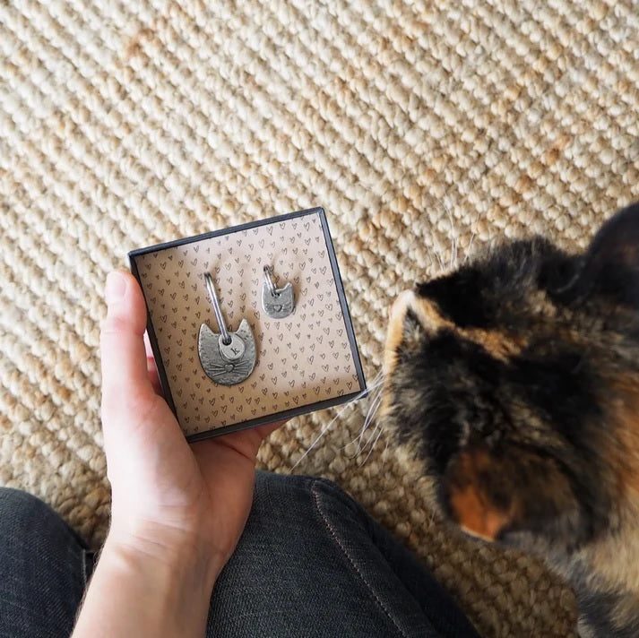 A person holding a box with two cat-shaped pewter keychain and charm, with a cat observing closely on a beige carpet.