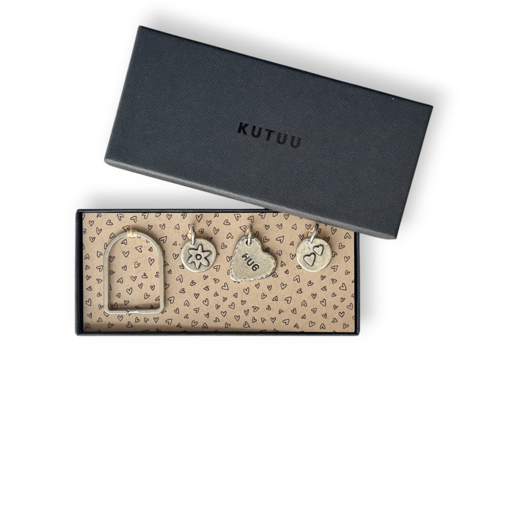 Black gift box with "KUTUU" on the lid, an arch-shaped keyring and three pewter charms on a heart-patterned lining.