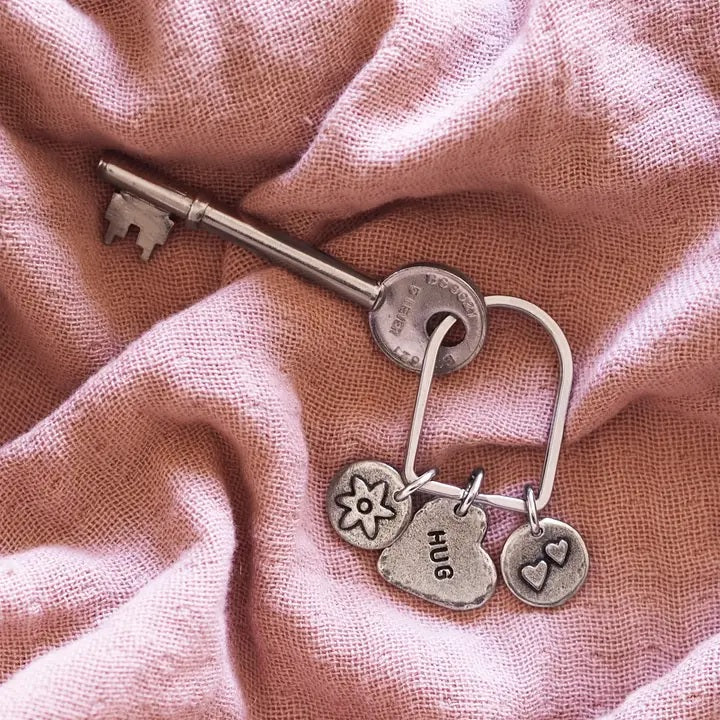 Silver key and a keyring with a heart-shaped "HUG" charm and other charms on pink fabric.