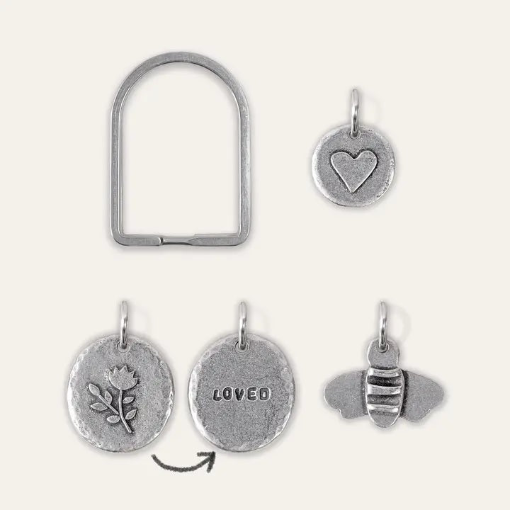 An arch-shaped keyring with three pewter charms in various designs, including a heart, a bee and a flower "LOVED" showing front and back designs.