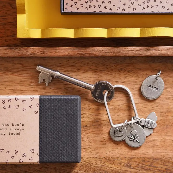 A silver key and a keyring with charms and a stamped "LOVED" charm on a wooden surface next to a heart-patterned package.