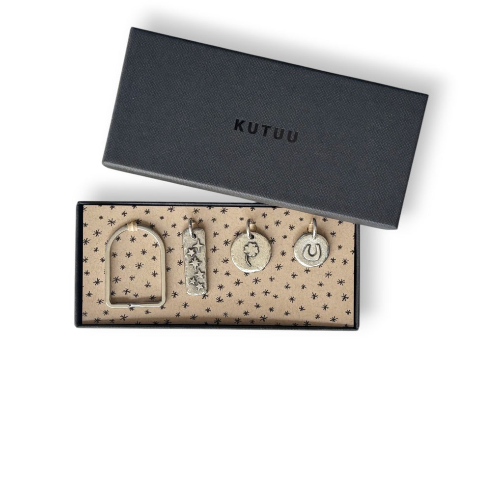 Black gift box with "KUTUU" on the lid, an arch-shaped keyring and three pewter charms on a star-patterned lining.