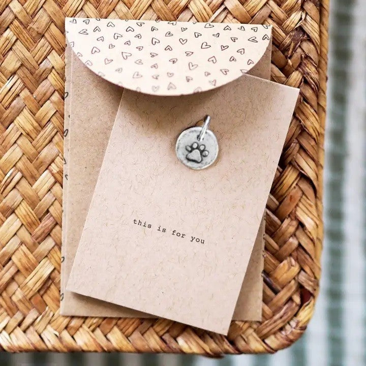 A kraft paper card with paw print embossed silver metal charm and envelope with hearts patterned flap on a woven tray.
