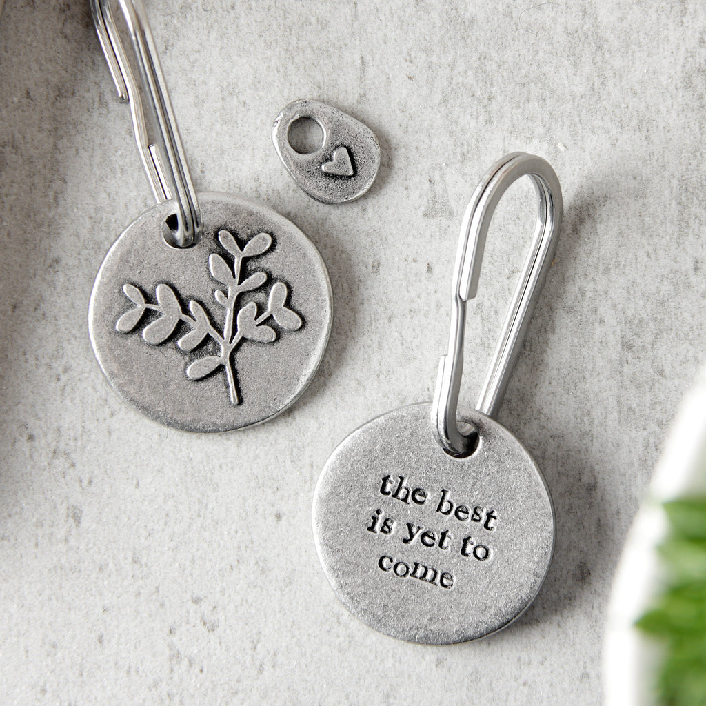 A round pewter keychain with a leaf embossed and engraved text details, showing front and back design on a textured surface.