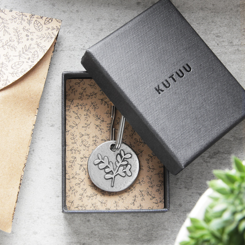 Gift box with a leaf design round pewter keychains on a leaves-patterned lining, with "KUTUU" printed on the lid, on a textured surface next to a matching envelope.
