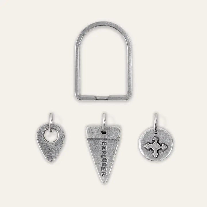 An arch-shaped keyring with three pewter charms in various designs, including a map pin, a triangular flag "EXPLORER", and a compass.