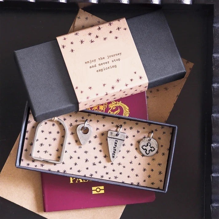 A gift box with an arch-shaped keyring and three pewter charms on a star-patterned lining, an inspirational message on the lid, on top of a red passport and an envelope.