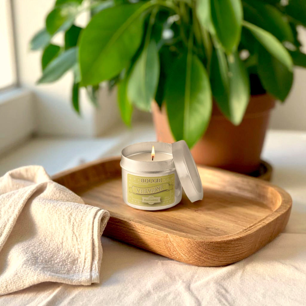 Maitre Savonitto verbena candle in an aluminum tin, lit, on a wooden tray near a plant.
