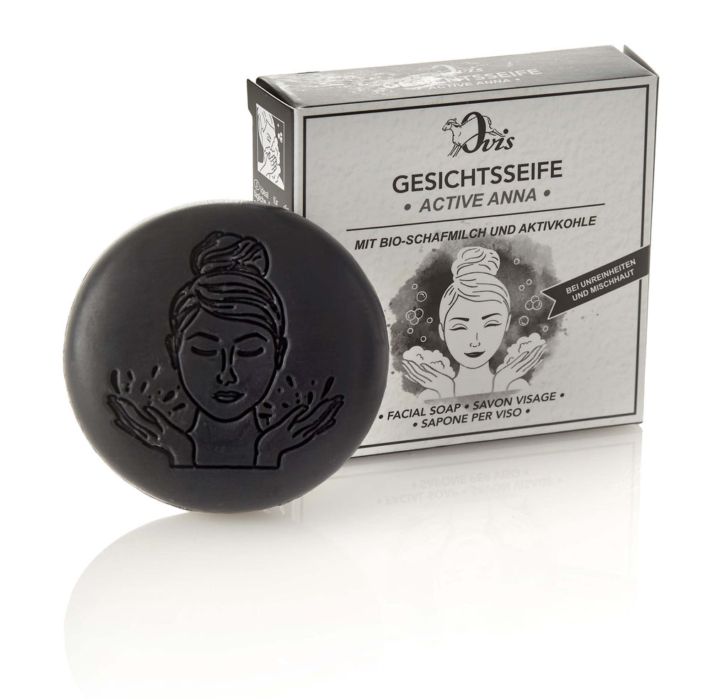 Round black facial soap with an embossed woman's face, positioned in front of a black and white box with matching illustration.