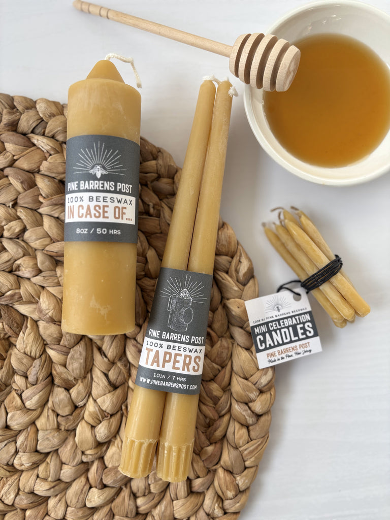 Various beeswax candles with brand name "Pine Barrens Post" and illustration and a bowl of honey with a honey dipper on a placemat.