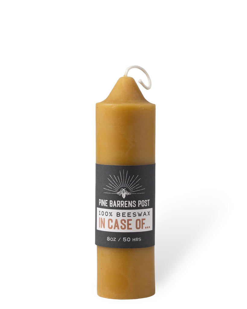 A beeswax pillar candle joined by a navy blue paper band with brand name "Pine Barrens Post" and illustration.
