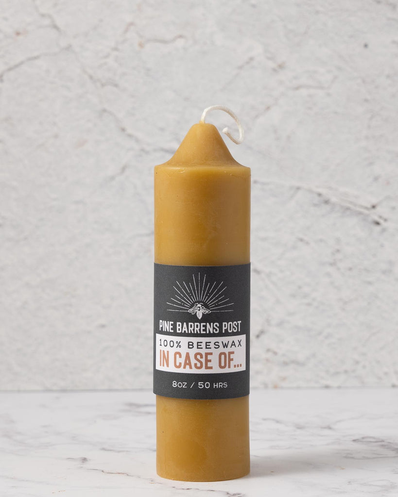 A beeswax pillar candle joined by a navy blue paper band with brand name "Pine Barrens Post" and illustration.