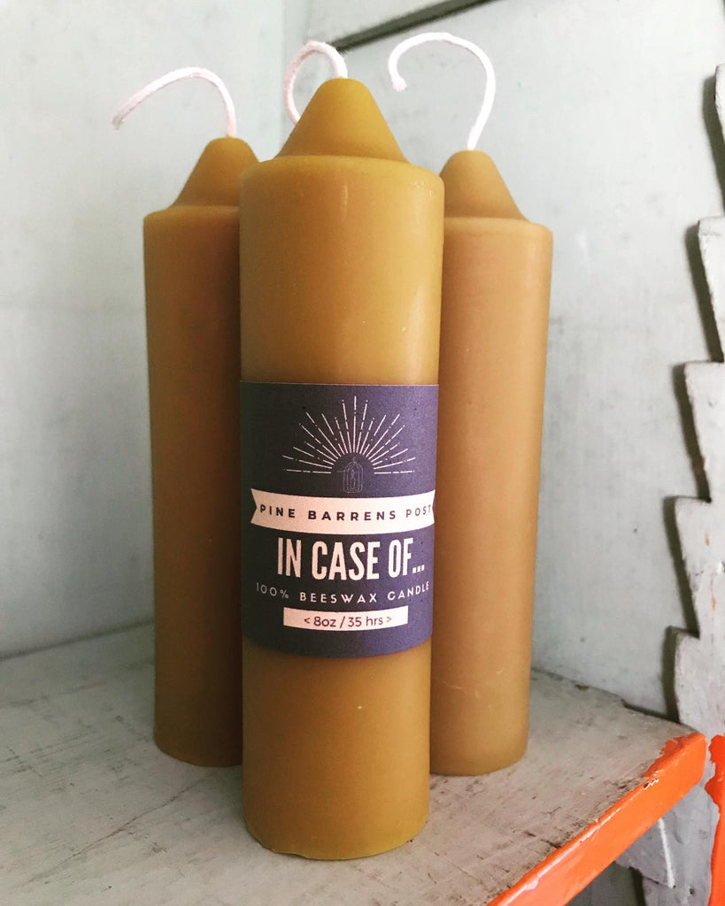 Three beeswax pillar candles, one wrapped with a navy blue paper band with brand name "Pine Barrens Post" and illustration.