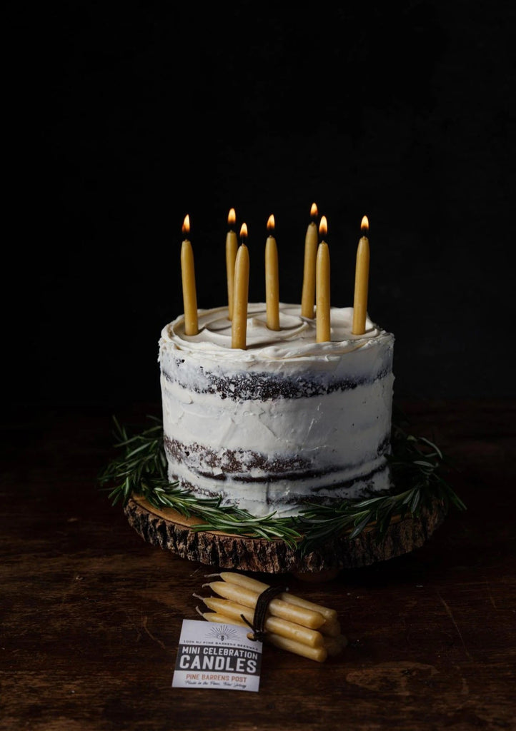 Rustic cake with lit candles, displayed on a wooden slab, surrounded by rosemary and a small bundle of "Pine Barrens Post" mini celebration candles in front.