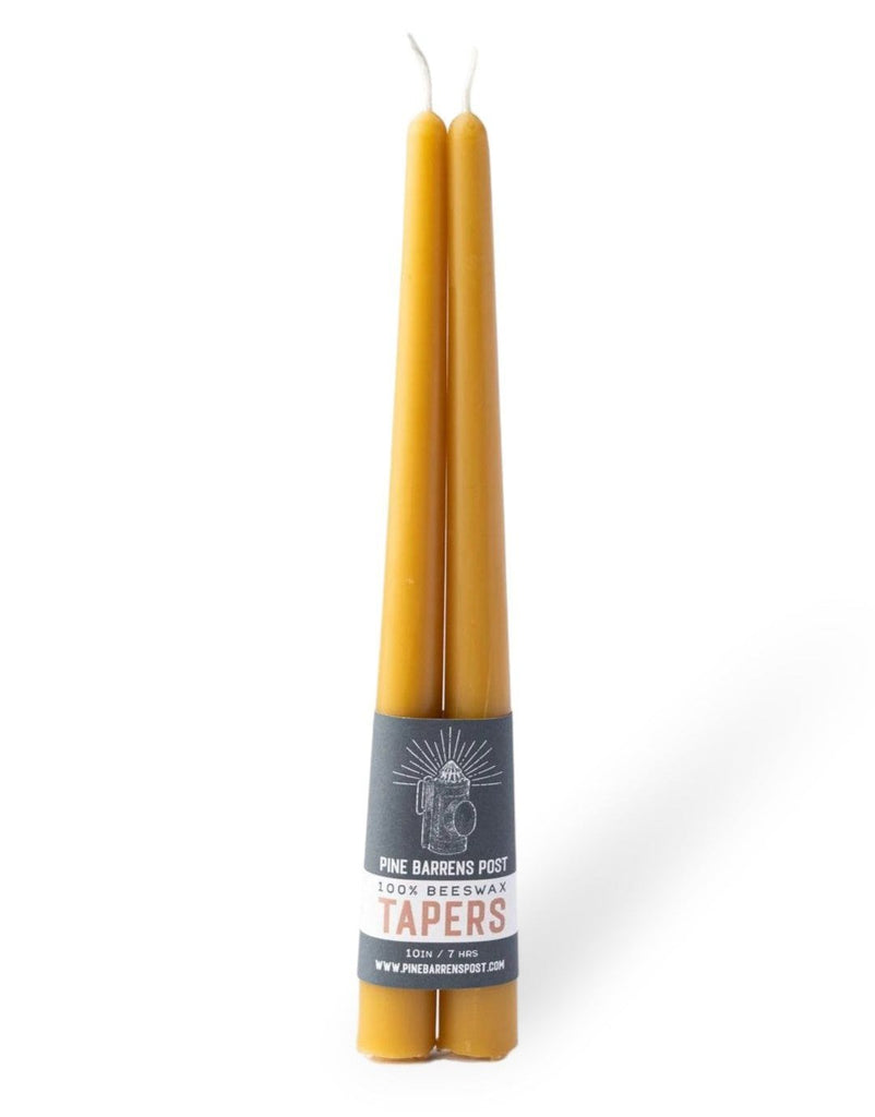 A pair of amber-orange beeswax taper candles joined by a navy blue paper band with brand name "Pine Barrens Post" and illustration.