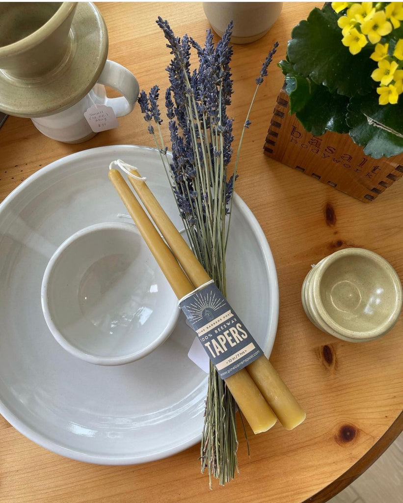 A tabletop display with a white plate and bowl, dried lavender, "Pine Barrens Post" taper candles, and a plant in a wooden box.