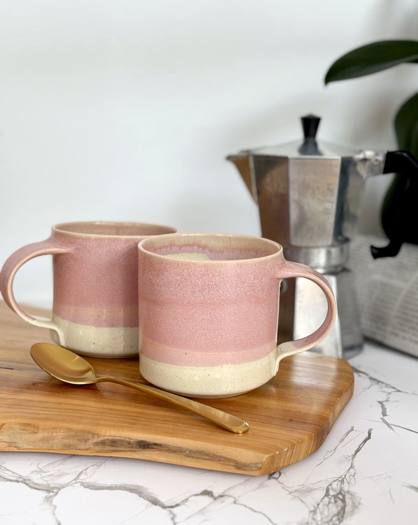 Two pink and beige two-toned gradient color ceramic mug on a wooden board with a gold spoon and an espresso maker in the background.