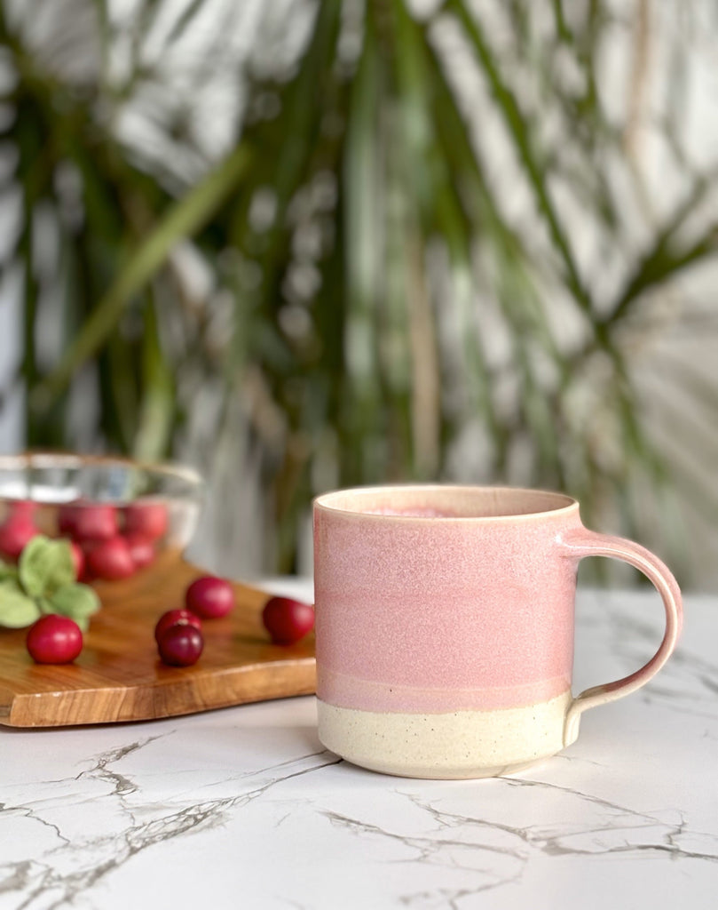 Two pink and beige two-toned gradient color ceramic mug on a marble surface with cranberries and palm plant in the background.