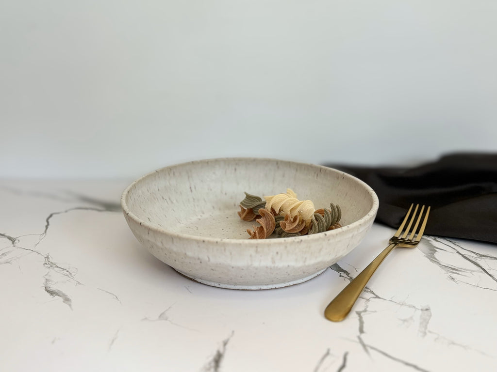 A beige speckled ceramic pasta bowl with spiral pasta on a marbled surface with a gold fork and dark napkin.