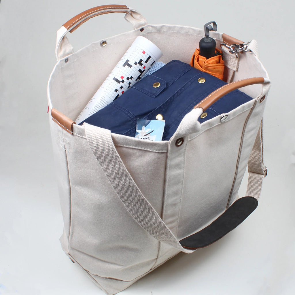 ShoreBags Natural Cargo Canvas Travel Tote bag filled with items including a crossword print paper, a navy shirt, and an orange umbrella.