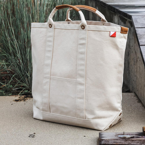 ShoreBags Natural Cargo Canvas Travel Tote bag with brown leather handles on a concrete surface next to a staircase and tall grass.
