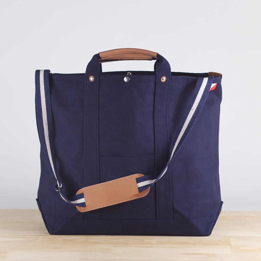ShoreBags Navy Cargo Canvas Travel Tote bag with brown leather handles and an adjustable shoulder strap on a light wooden surface.
