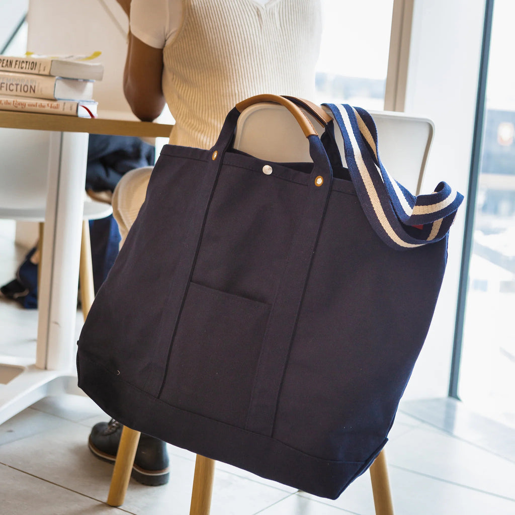 ShoreBags Navy Cargo Canvas Travel Tote bag with leather handles and a blue and white striped strap hanging on a chair.