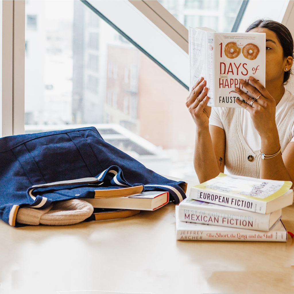 Person reading a book at a table with a stack of books and a ShoreBags Navy Cargo Canvas Travel Tote bag on the side.
