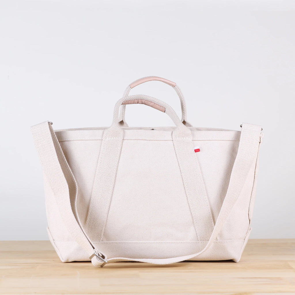 ShoreBags Utility Heavyweight Canvas Tote bag with handles and an adjustable shoulder strap, featuring a red tag on a light wooden surface.