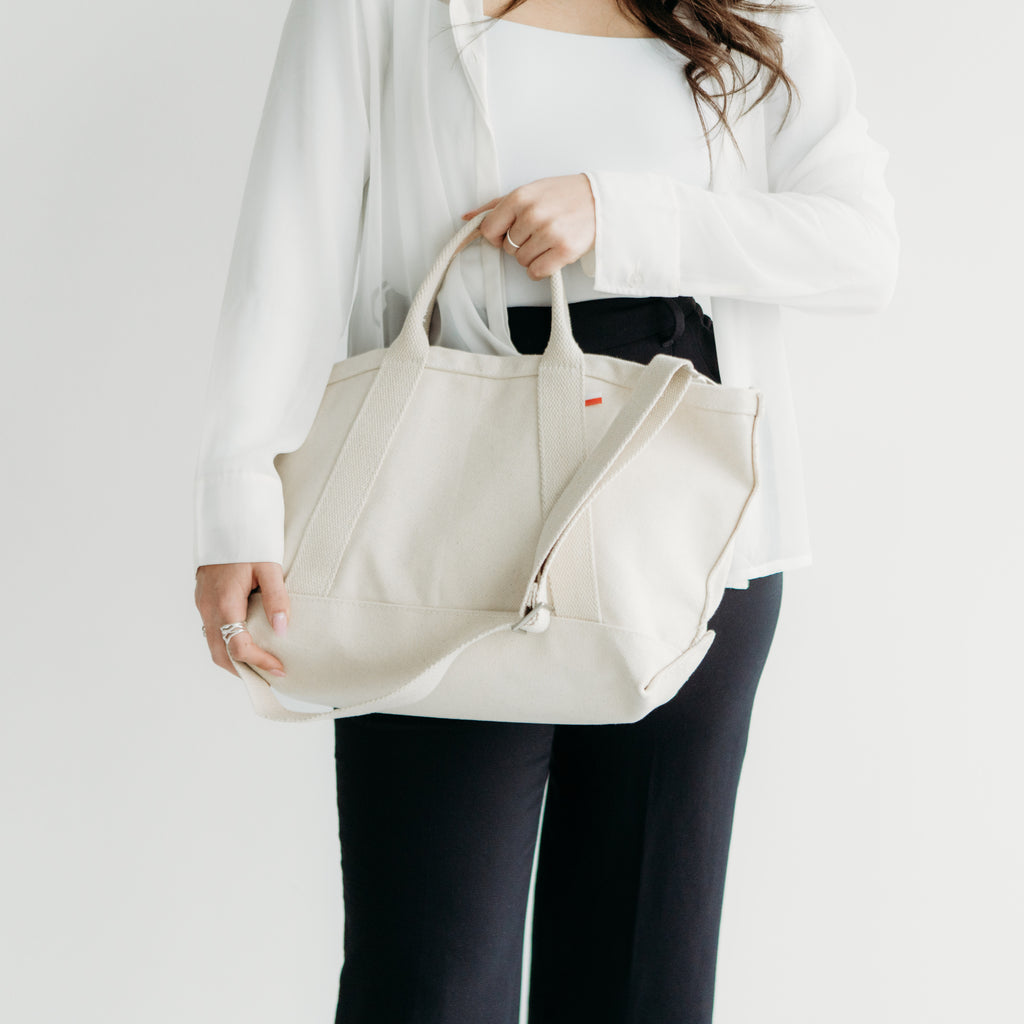 A person holding a ShoreBags Utility Heavyweight Canvas Tote bag with handles and an adjustable shoulder strap.