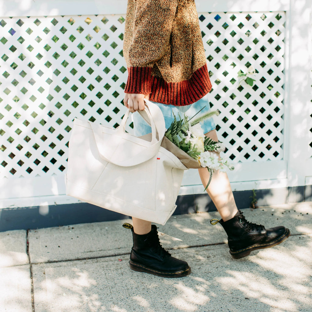 Person in a sweater and shorts walking with a ShoreBags Utility Heavyweight Canvas Tote bag and flowers, wearing black boots, near a white lattice fence.