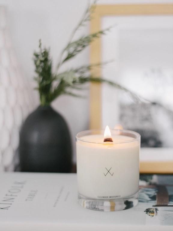 A lit candle labeled "Cucumber Sweetgrass" with a vase and framed picture in the background.