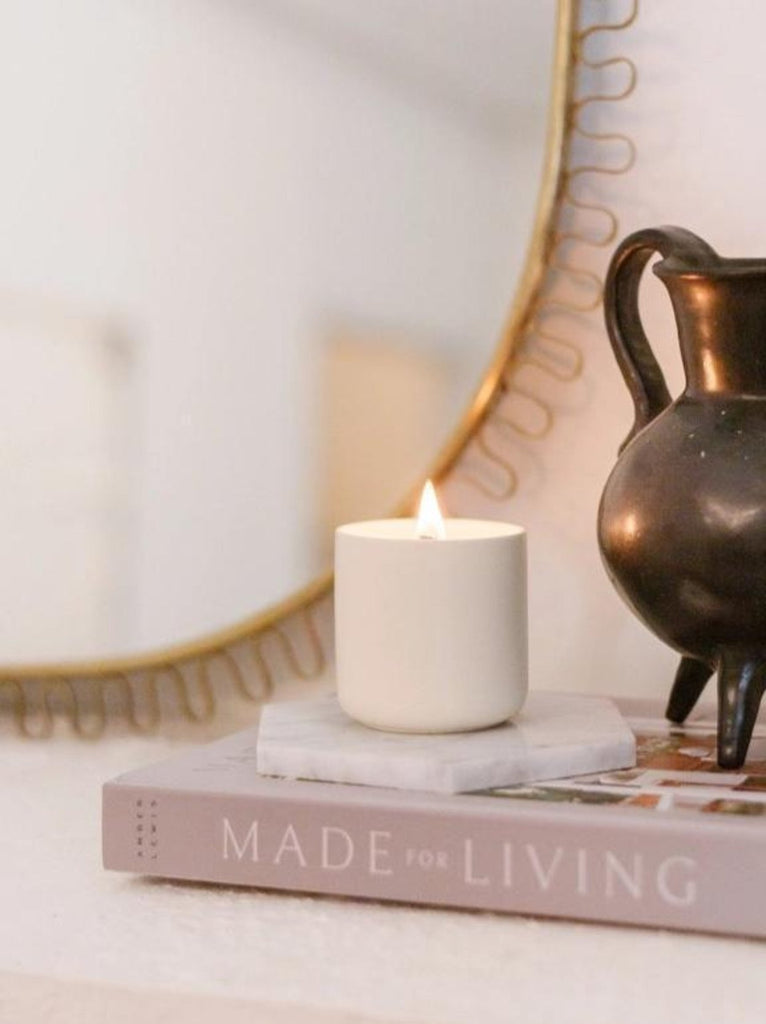 A lit candle on a hexagon marble coaster, next to a bronze-toned pitcher, on a book titled "MADE FOR LIVING" on a table, with a decorative mirror in the background.