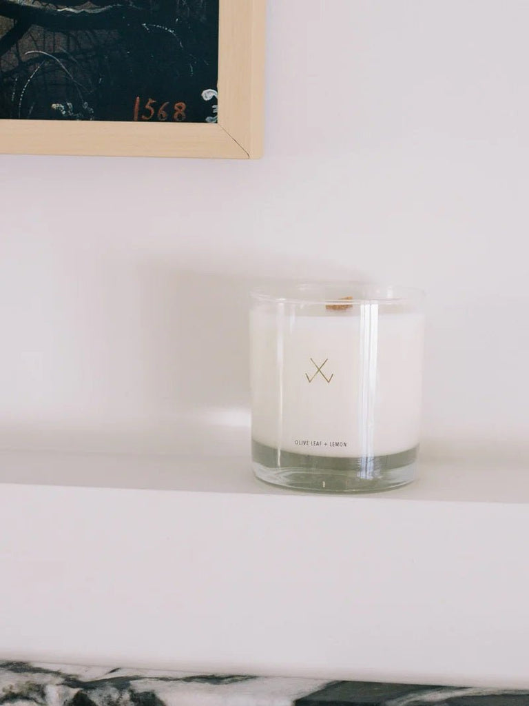A glass candle labeled "Olive Leaf + Lemon" on a white shelf with a framed picture above.