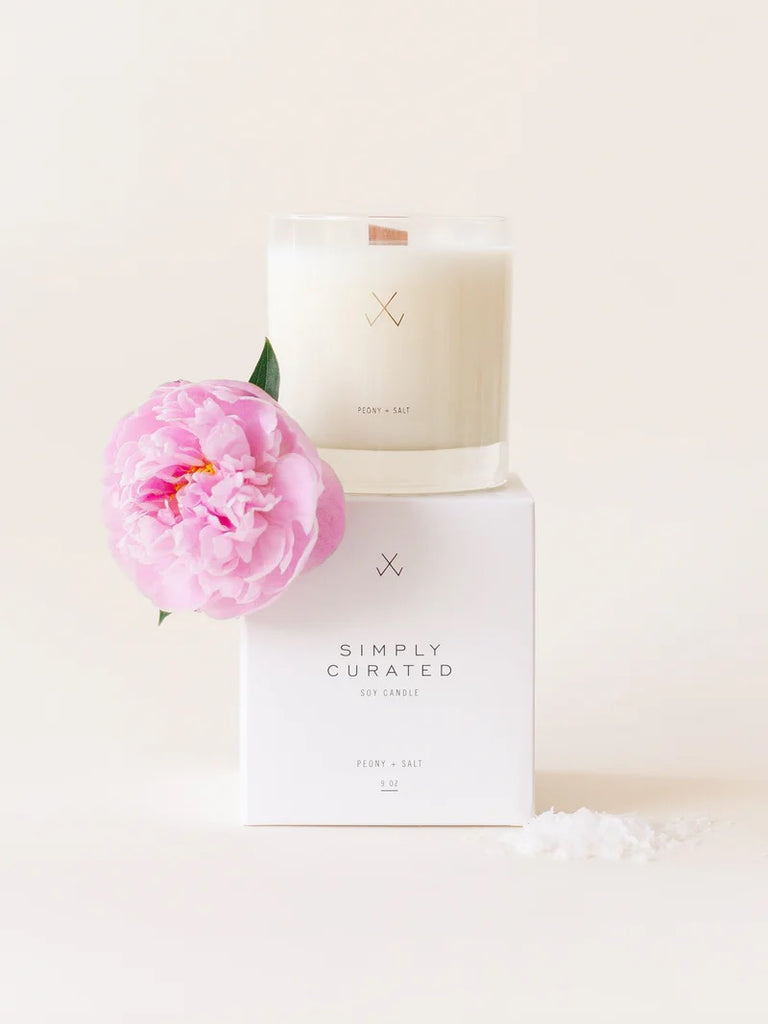 A Simply Curated peony + salt soy candle on its white packaging box with minimalistic branding logo, with a pink peony flower and a pinch of salt.