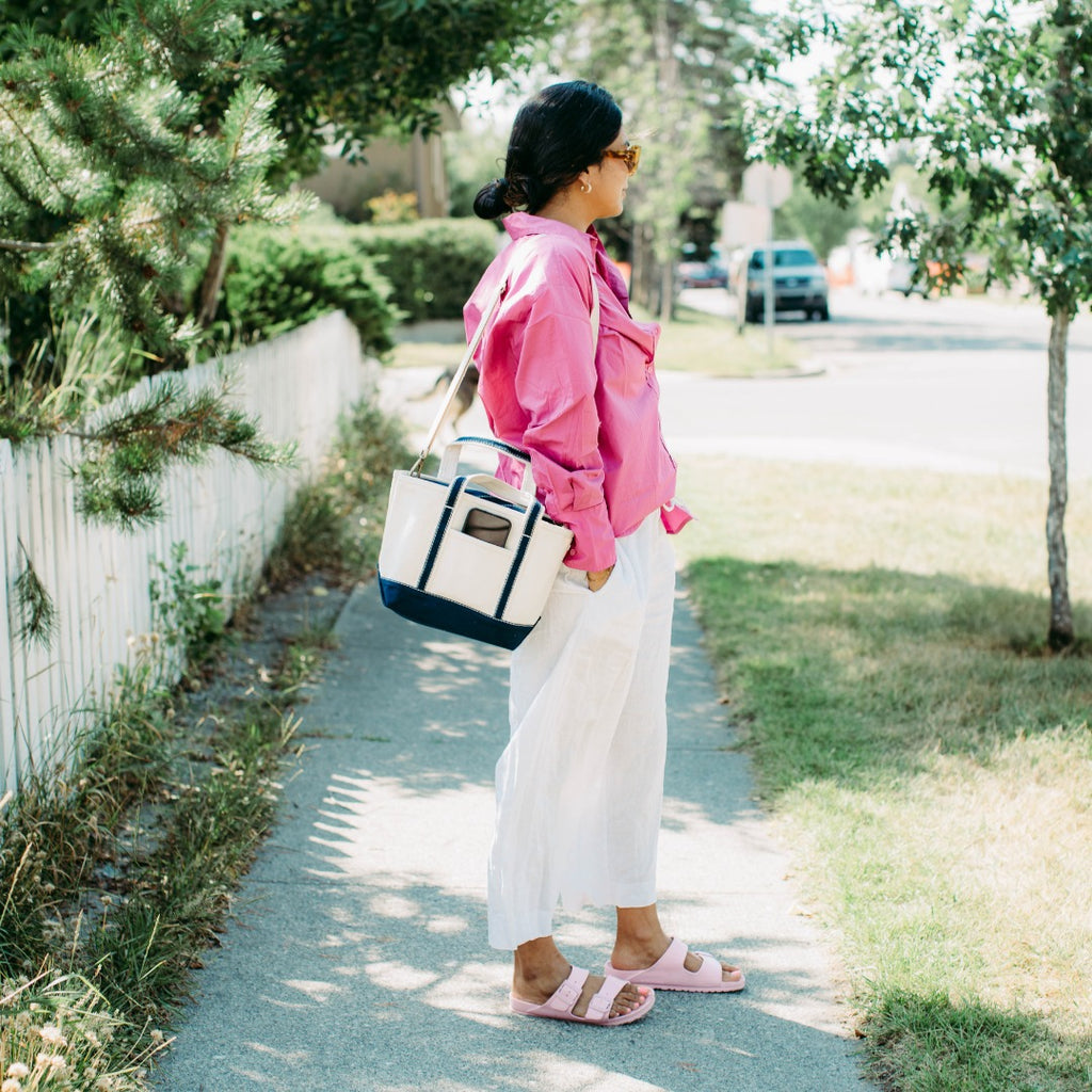 Person in pink shirt and white pants standing on a sidewalk with ShoreBags Sonoma Canvas Crossbody Tote bag.