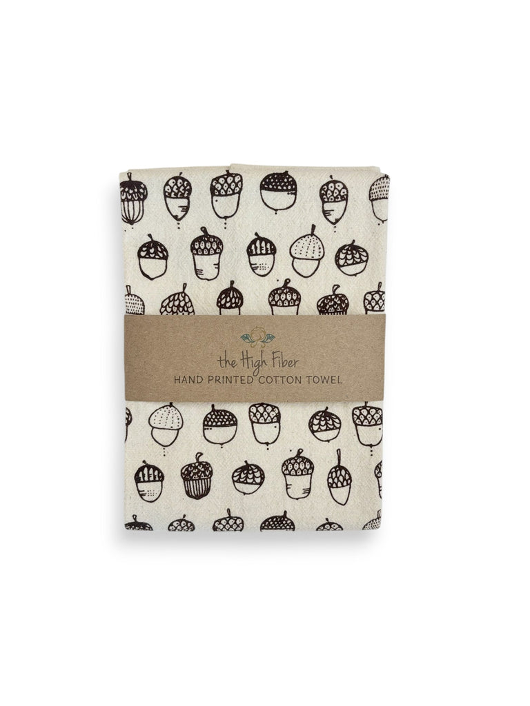 A folded natural cotton tea towel with brown acorns patterns and a brown paper band with brand name "the High Fiber" and logo.