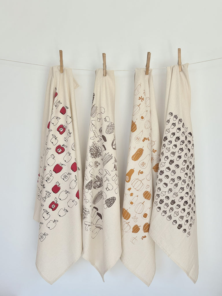 Four natural cotton tea towels with apple, mushroom, winter squash, and acorn patterns hanging on a clothesline.