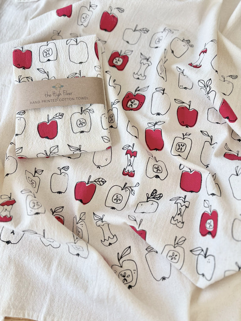 A natural cotton tea towels with red and black-outlined apples patterns, with another tea towel wrapped in a brown paper band with "the High Fiber" and the logo printed on it. 