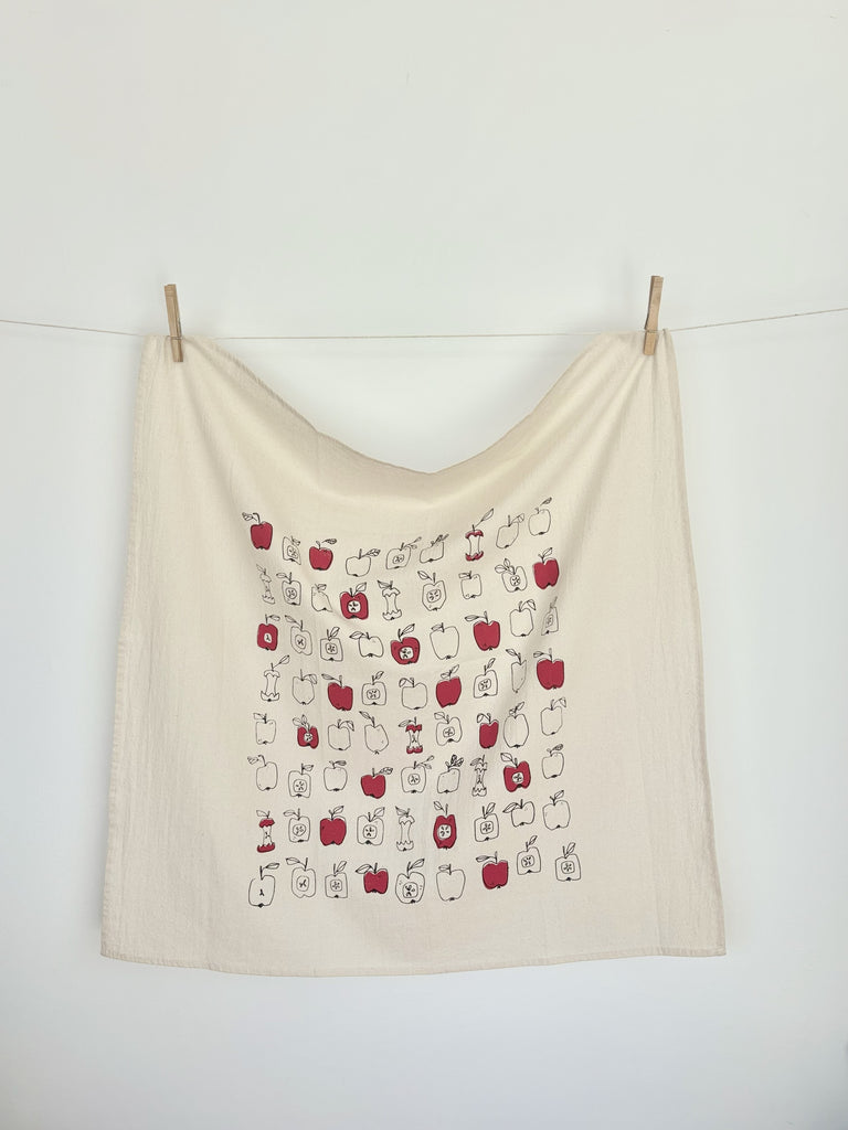A natural cotton tea towel with red and black-outlined apples patterns hanging on a clothesline.