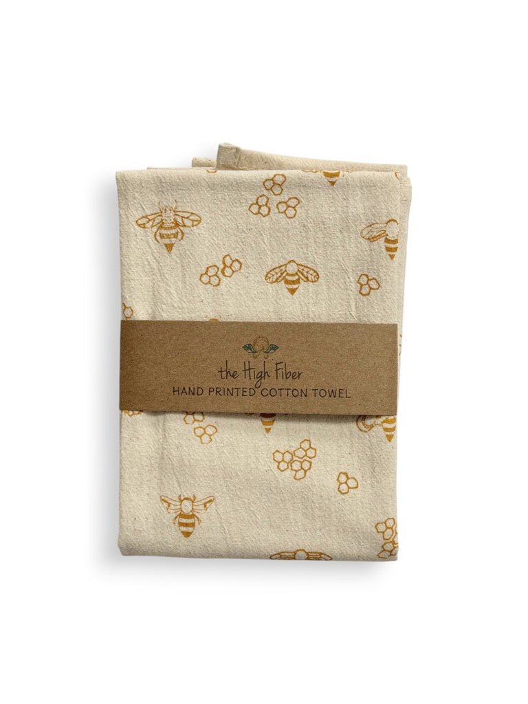 A folded beige tea towel with gold honeybees and honeycombs patterns and a brown paper band with brand name "the High Fiber" and logo.