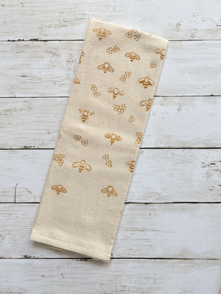 Beige tea towel with gold honeybees and honeycombs patterns on whitewashed wood surface.