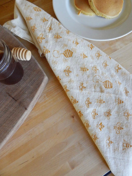 A light wooden table with honey, a honey dipper, a plate of pancakes, and a honeybees-patterned tea towel.