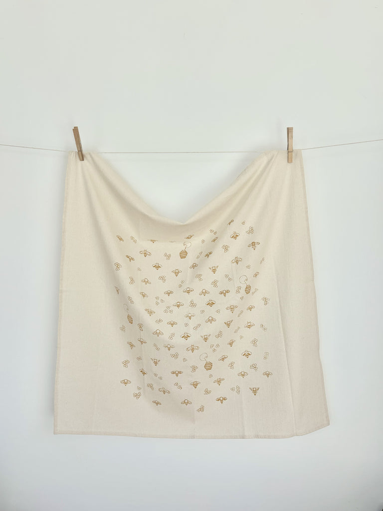 A natural cotton tea towel with gold honeybees, honey pots and honeycombs patterns hanging on a clothesline.